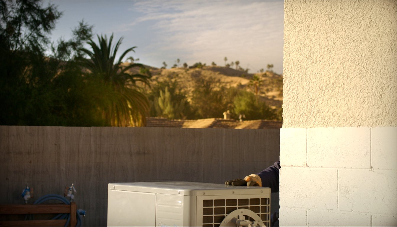 Outdoor AC condenser at a residential home in Jurupa Valley, California with palm trees and Santa Ana River corridor backdrop, gloved JC Energy Solutions technician hand on the service valve