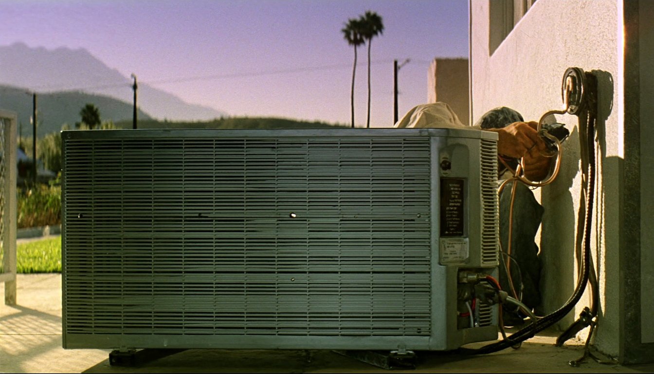 Outdoor AC condenser at a residential home in Rialto, California with palm trees and northern San Bernardino mountains backdrop, gloved JC Energy Solutions technician hand on the service valve
